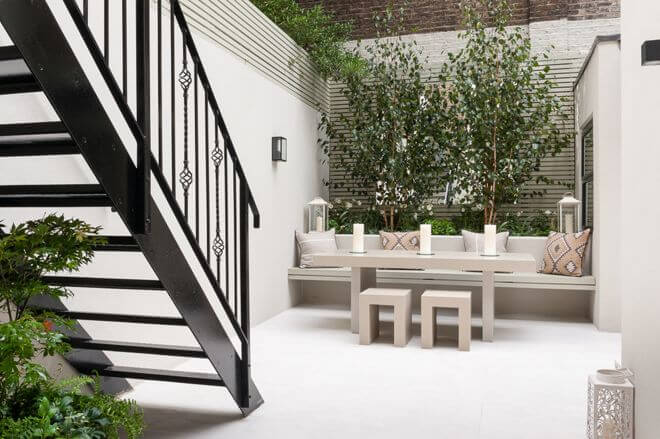 Consider built-ins. Obviously, space is at a premium with a small patio. In this modest courtyard, the raised garden bed serves as the back of a built-in bench. This adds room for a generously sized table in the space. Tall trees and slatted trellis walls keep the building walls around the courtyard from feeling oppressive.
