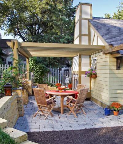 Patios typically are located off the house, but if you don’t have room to expand there, a detached garage or shed can provide a nice anchor for an outdoor living space. Here, a wood-burning fireplace and window boxes off the garage add warmth and charm to the patio, and the structure also provides a place to anchor a pergola.